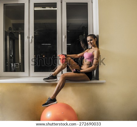 athletic young beautiful woman with protein cocktail in plastic shaker sit on windowsill Leg stand on fitness ball on wall texture background. Sports nutrition. Resting after powerful exercise in gym