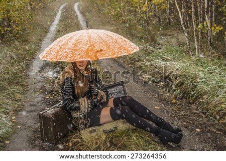 No face Unrecognizable person girl under orange snowy umbrella, late autumn Adult woman sit in open old retro vintage aged suitcase on land way road between green and yellow grass field with trees