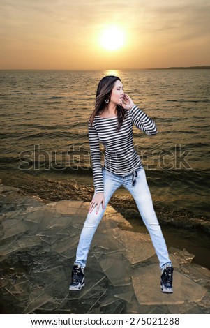 Full length Beautiful sexy brunette woman wear sailor striped dress and blue sexy jeans posing near sea water texture Asian girl stand on debris of ice on sand coast against sunset sky with sun