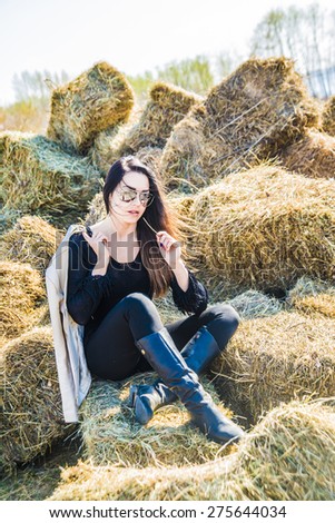 portrait of beautiful girl with long hair sit on hay block, cute woman rest in field against spring sky Female wear black stylish dress and leather high boots Sunny day on fly White jacket on shoulder