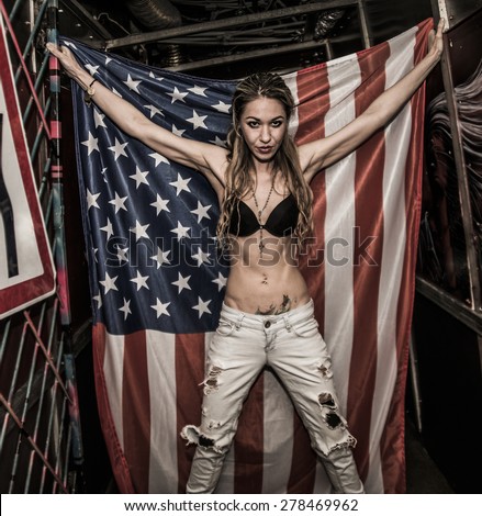 Full length image of a sexy slim blond woman hold in hands big American flag Skinny body of cute young adult girl with black pantyhose on legs stand in corridor perspective in dark background