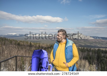 Man stand near big backpack on a cliff looking at snowy mountain forest Handsome male against blue sky background Heaven with clouds Sunny day Windy cold weather Empty space for inscription