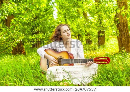 Woman playing acoustic guitar in central park Cute young adult girl sit on fresh green grass against trees in perspective  Summer meadow or field Spring time Female wear white dress and skirt