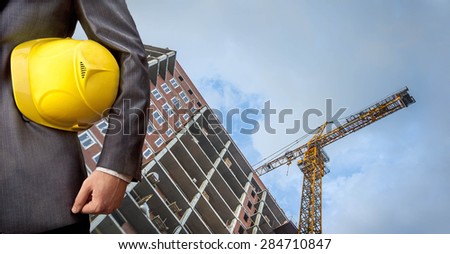engineer holding yellow helmet for workers security on background of new highrise apartment buildings and construction cranes on background of evening sunset cloudy sky Silhouette Crane lifts load
