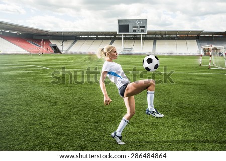 Young adult girl playing football or soccer, kicking a ball with her knee Beautiful blond woman in a sports suit stand on her legs on a green grass football field against empty stadium and scoreboard