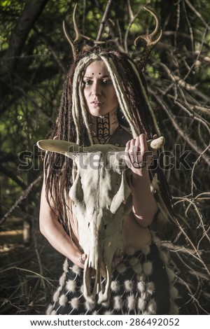 portrait of Beautiful woman with long dreadlock hair hold in hands cow skull with horns against  wild forest trees Young girl look at camera Woman shaman in ritual garment stand from fur and leather