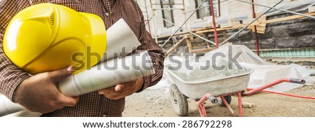 engineer hold blue print, yellow helmet for workers security on background of new apartment buildings and construction scaffolding and metal trolley on background Paper plans in document