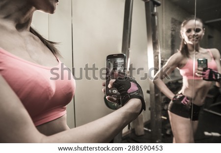 portrait of young adult woman with smartphone taking mirror reflection selfie in gym sport, fitness, lifestyle, technology and people concept Image of small screen of mobile cell phone Dusk light