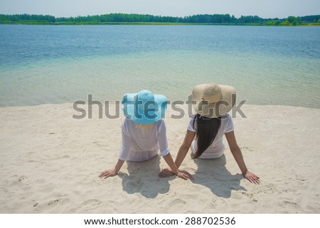 Lesbian Couple in white clothes sit near each other on a tropical white sand beach at island Brunette and blond woman look far away on water texture Two cute girl against blue sky with clouds