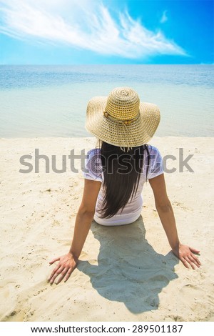 Summer vacation woman sitting on yellow sand coast wear beach hat enjoying summer holidays looking at water texture in ocean. Beautiful back side of model Cute girl against blue sky with clouds