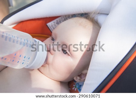 mother feeding her naked baby son with milk from plastic bottle in stroller outdoor at summer park Empty space for inscription