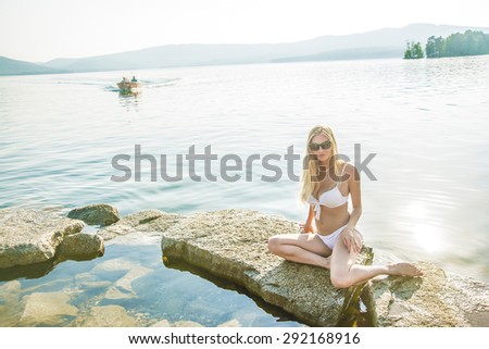 Girl at the sea. Attractive young blond woman in white swimsuit posing at the sea The sexual young blonde girl with a beautiful body sunbathes on a beach in a white bathing suit against  sea