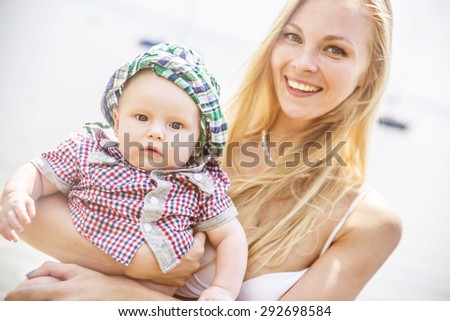 Beautiful baby on a beach with his young blond mother looking at camera Cute blondy mom Caucasian woman wear white swimsuit against water sea background Summer family travel on coast