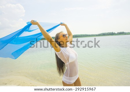 Beautiful young adult girl holding blue fabric at wind in a summer vacation Cute woman wear white transparent clothes and orange sunglasses against water texture and blue sky with clouds