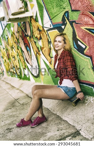 Portrait of beautiful teen girl sitting on skateboard over wall background with graffiti art. Urban outdoors, teenager\'s lifestyle. Full length Cute young adult woman sit skate in denim jeans shorts