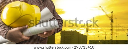 engineer hold yellow helmet for worker security blue print paper document on background of highrise apartment buildings and construction cranes on background of sunset sky Silhouette Crane lifts load