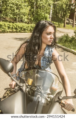 Portrait of  beautiful brunette woman sit on black sports motorcycle Very hot sexy young femme fatale girl wear denim vest.  Sexy Lady against green summer bush background