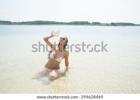 Beach holiday. A smiling brunette hair woman staying at water and pours water from a large conch shell onto herself chest Happy sexy young woman pouring liquid from the big seashell against blue sky