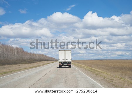 Alone Dirty Truck driving on clear spring asphalt  road against blue sky with white clouds, empty field with dry grass and forest along highway Back view car Idea symbol of logistics