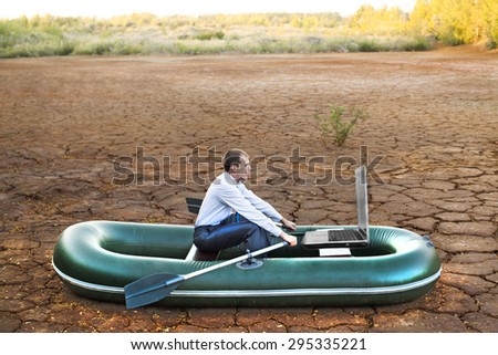 Business man will rows home for shore in paddle powered row boat in boat looking at  laptop rocks looks future symbol crisis losses braking difficulties environmental disaster water scarcity drought