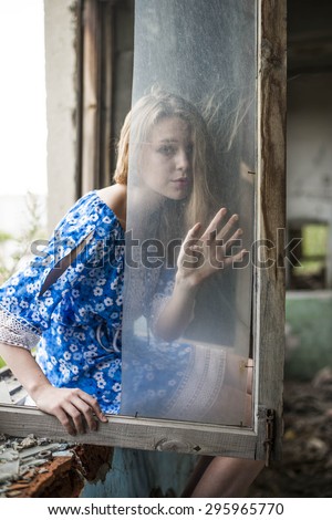Full length portrait of Young adult woman sit on windowsill near dirty glass window. Girl\'s hand lie on dirt transparent texture Windy day Hair fly in air Female wear blue dress with flowers pattern