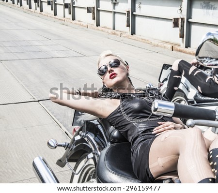 Biker Woman on motorcycle against metal bridge background Young brutal cute girl lie on chopper and wear black leather dress and stylish sunglasses  Empty space for inscription