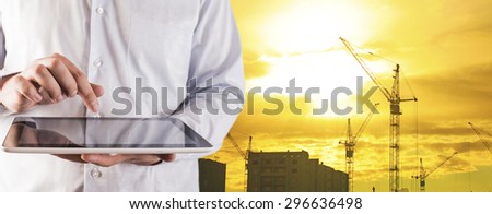 Engineer and architect hold digital tablet computer on sunset sky with clouds and sun background No face Unrecognizable person New highrise apartment buildings and construction cranes Silhouette Crane