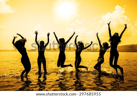 Silhouette of young group of people jumping in ocean at sunset Team of adult girl stand in water on summer beach against blue sky with clouds Water splash Hair fly in air Empty space for inscription