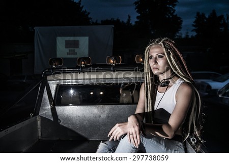 Sexy freak style woman sitting in metal car body of retro pick up auto Cute woman with dreadlocks smoke cigarette against dark night summer forest Female with headphones
