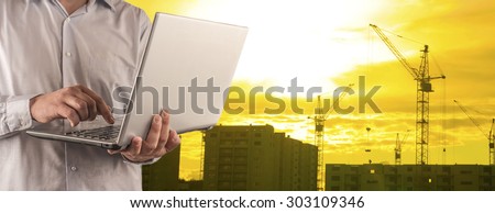 Engineer and architect hold digital laptop computer on sunset sky with clouds and sun background No face Unrecognizable person New highrise apartment buildings and construction cranes Silhouette Crane