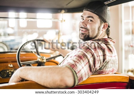 man looking at auto through old retro vintage aged car window Hand lie on wheel Beard Gay wear protective glasses from leather and glass goggles and cap Near door weighs spare wheel