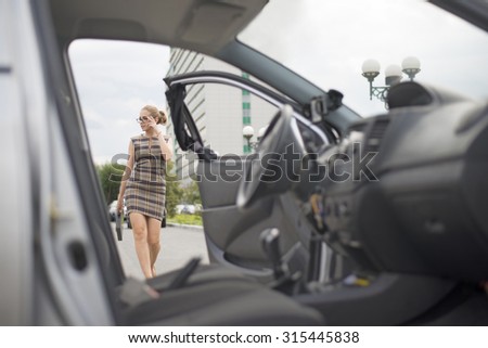 Getting ready. Portrait of young smiling serious lady going to car with opening door. Empty auto interior inside. Stylish Businesswoman wear short dress and glasses with plastic case