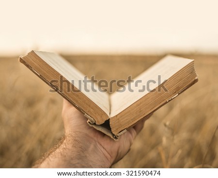 male hands holding opened hardback book, diary with fanned pages on blurred nature landscape background against wheat field and sunset sky back to school education concept.