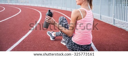 No face. Cute slim young adult girl hold in hand black plastic shaker. Blonde woman sitting on running track and drinking bottled water. Empty sporty space for inscription or other objects and people