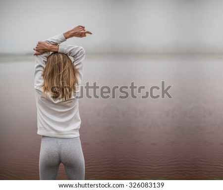 Side rear view of a young sporty woman stretching her arms up while standing on a beach by the shore and against a gray sky. Dirty red toxic water in lake. ecological catastrophe