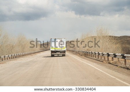 Alone clean Truck driving on clear spring asphalt road against blue sky with white clouds, empty mountain horizon with dry trees and forest along highway Back view car Idea symbol of logistics