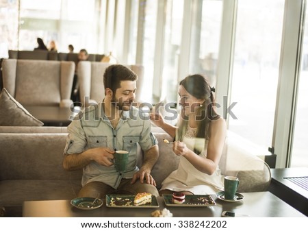 Romantic young couple drink coffee with handsome man laughingly eat biscuit to his girl friend or wife Cute woman look at cake Near together against glass window view in perspective Sweet food on fork