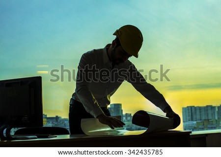 Silhouette builder engineer wear security helmet look at blueprint paper construction drawing plan on background of sunset window frame blue yellow sky with clouds near pc monitor computer
