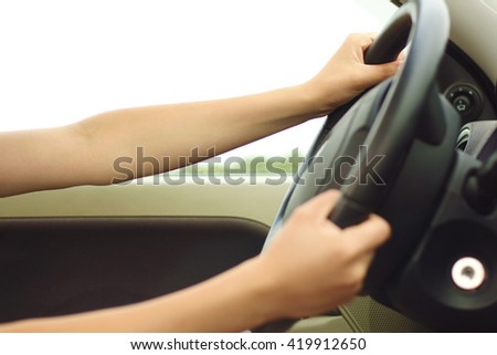 Close up of woman\'s hands driving a car. Picture of girl\'s arms holding steering wheel inside auto. Young female sitting indoor on summer outdoor background. no face