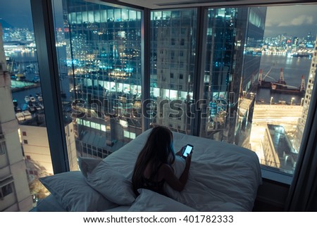 Young girl or teenager with long hair girl checking her mobile phone or chatting with someone at night in her bed with breathtaking view over night city. Hong Kong, China.