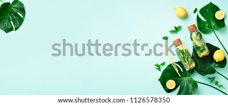 Bottle of detox water with mint, lemon and tropical monstera leaves on blue background. Flat lay. Banner. Citrus lemonade. Summer fruit infused water. Top view with copy space