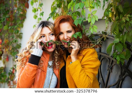 Outdoor lifestyle fashion portrait of two pretty cheerful girls friends, smiling and fooling around. Walking on the autumn city. Wearing stylish bright outerwear with handbags and sunglasses.