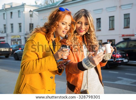 Outdoor lifestyle fashion portrait of two pretty cheerful girls friends, looking at the mobile phone. Smiling and drinking coffee. Chatting with friends. Wearing stylish bright outerwear