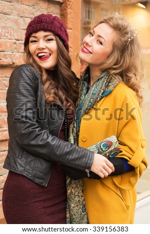 Outdoors lifestyle fashion portrait of two pretty cheerful girls friends. Smiling and walking on the autumn city. Wearing stylish outerwear, hat and sunglasses. Close up. Autumn colors. Joy, fun