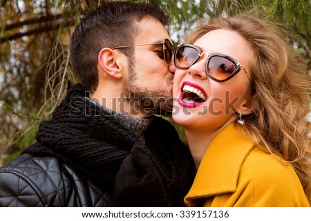 Close up portrait of happy smiling couple in love, guy kissing his girlfriend. Wearing bright outerwear, sunglasses. Outdoors lifestyle fashion portrait. Handsome brunet with beard and stunning blonde