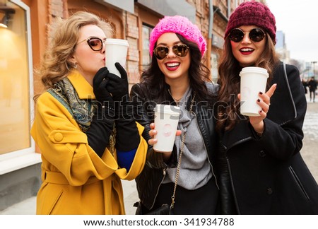 Outdoors fashion portrait of three young beautiful women friends drinking coffee. Smiling and going shopping. Kissing a cup of coffee. Wearing stylish outerwear, hats and sunglasses. Bright make up
