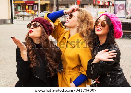 Outdoor lifestyle fashion portrait of three pretty cheerful girls friends, smiling and having  fun. Waving to someone. Walking on the autumn city. Wearing stylish bright outerwear, hats and sunglasses
