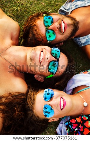 Outdoors lifestyle portrait of company young pretty best friends. Smiling, lying on the grass and looking at the sky. Wearing bright t-shorts and stylish sunglasses. In sunglasses reflected palm trees
