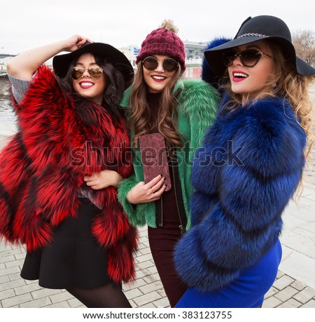 Outdoors lifestyle fashion portrait of company pretty cheerful girls best friends in stylish hats and sunglasses. Wearing bright colored fur coats. Smiling and walking on the city. Close up