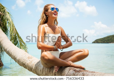 Summer lifestyle portrait of pretty happy young girl with tanned sexy body. Doing yoga, smiling and sitting on palm tree at the tropical island beach with clear water. Wearing stylish bikini, sunglass